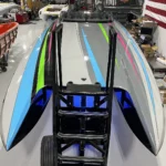 Custom high-performance boat showcasing sleek design with vibrant blue, green, and pink stripes, positioned on a cart in a marine workshop environment.