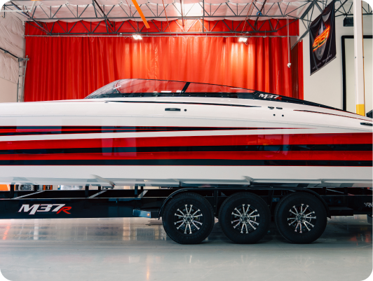 Custom high-performance boat model M37R displayed on a trailer, showcasing sleek design with red and white stripes, in a well-lit manufacturing facility.