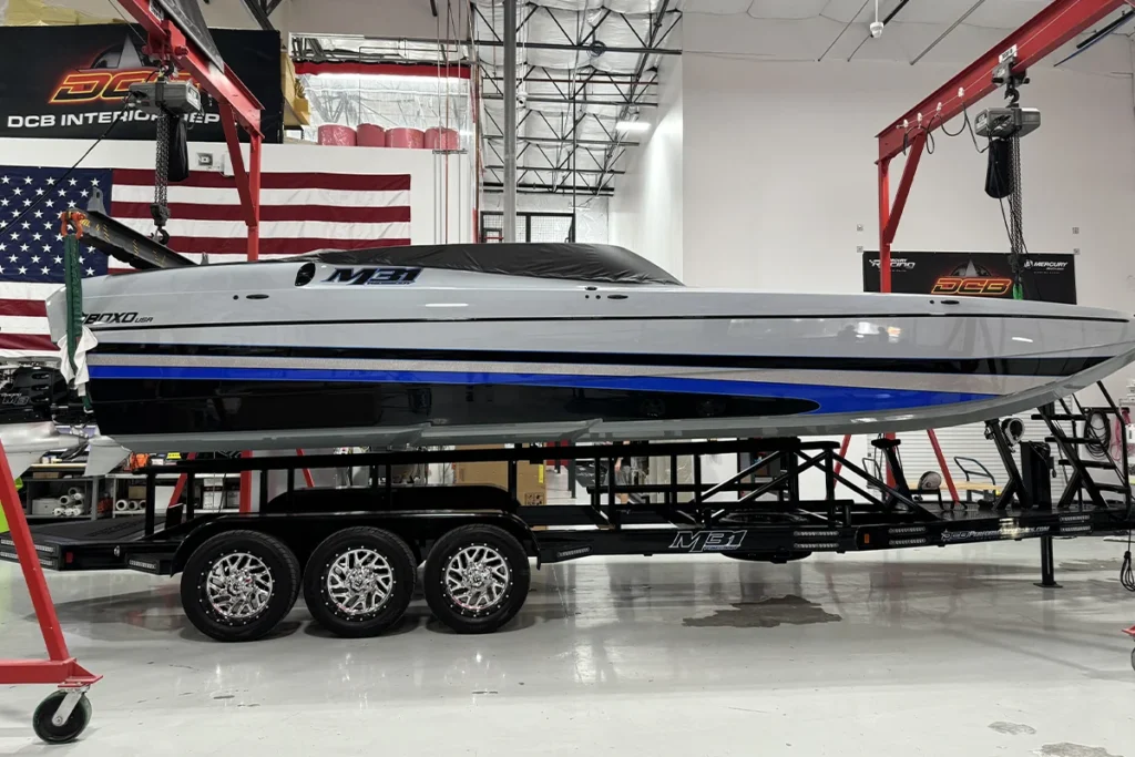 DCB Performance Marine custom boat on trailer, showcasing sleek design with blue and black accents, suspended in factory setting with American flag in background, highlighting craftsmanship and attention to detail in the build process.